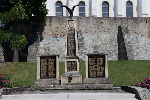Ein Denkmal mit drei Steinsäulen, die jeweils mit Namen graviert sind, steht vor einer Steinmauer mit Bogenfenstern. Auf der mittleren Säule thront eine Adlerstatue.