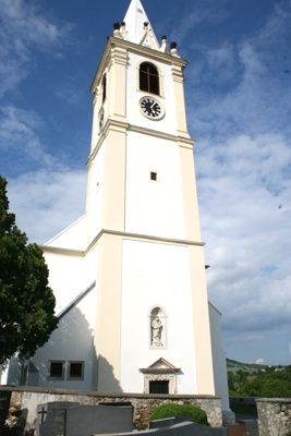 Ein hoher weißer Kirchturm mit einer Uhr, vor einem blauen Himmel mit Wolken.