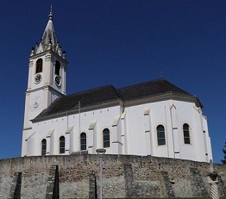 Eine große weiße Kirche mit einem Turm und einem Uhrenturm steht gegen einen blauen Himmel, umgeben von einer Steinmauer.
