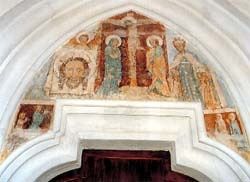 Ein Fresko mit religiösen Figuren, darunter Jesus am Kreuz, Maria und Heilige, schmückt den Bogen über einer Tür in einer Kirche.