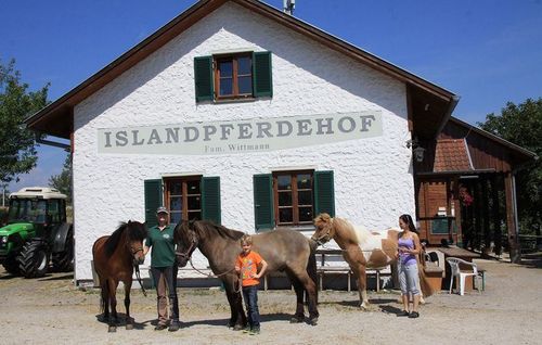 Drei Personen und ein Kind stehen vor einem weißen Gebäude mit Pferden, einer hält die Führstricke eines Pferdes. Das Gebäude trägt das Schild 'ISLANDPFERDEHOF Fam. Wittmann'. Ein Traktor ist links geparkt.