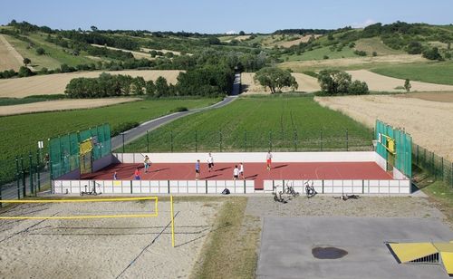 Eine Draufsicht auf ein Sportfeld mit Menschen, die Basketball und Volleyball spielen. Das Feld ist von grünen Feldern und Bäumen umgeben.