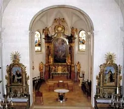 Das Innere einer Kirche mit einem Hauptaltar, der ein großes Gemälde zeigt, flankiert von Statuen und geschmückt mit aufwendigen Dekorationen, einschließlich eines goldenen Sonnenstrahls und kunstvollen Rahmen.