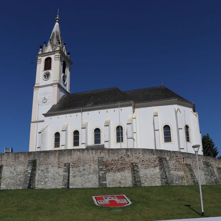 Eine weiße Kirche mit einem schwarzen Dach und einem hohen Turm mit einer Uhr. Die Kirche ist von einer Steinmauer umgeben. Im Vordergrund befindet sich ein Rasenbereich mit einem roten und weißen Logo.