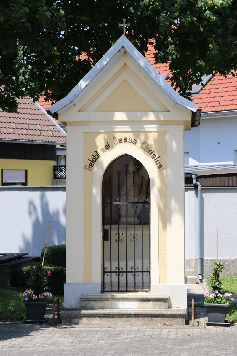 Eine kleine Kapelle mit einem schwarzen Metalltor und einer Statue von Jesus Christus im Inneren, in einem Garten mit rosa Blumen und grünen Büschen gelegen.