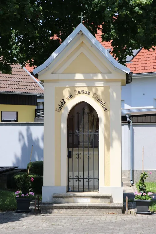 Eine kleine Kapelle mit einem schwarzen Metalltor und einer Statue von Jesus Christus im Inneren, in einem Garten mit rosa Blumen und grünen Büschen gelegen.