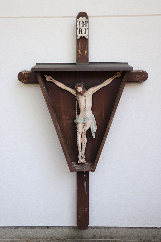 Ein hölzernes Kruzifix an einer Wand mit einem dreieckigen Rahmen und der Aufschrift 'IN RI' darüber. Das Kruzifix zeigt eine Figur von Jesus Christus am Kreuz, der ein Lendenschurz und eine Dornenkrone trägt, mit einem Rosenkranz an seiner Seite.