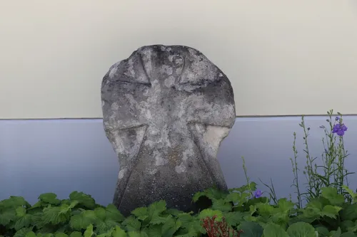 Ein altes Steinkreuz steht in einem Garten, verwittert und mit Moos und Flechten bedeckt.