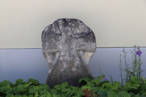 Ein altes Steinkreuz steht in einem Garten, verwittert und mit Moos und Flechten bedeckt.