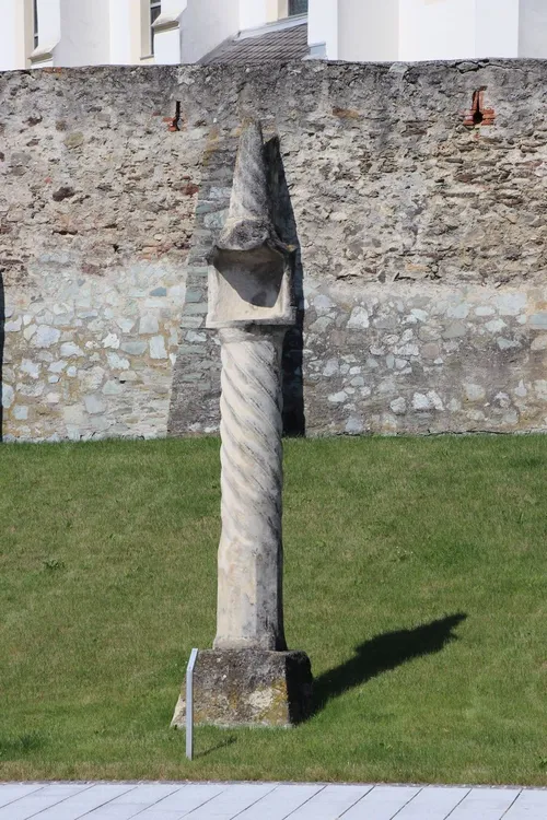 Eine alte Steinsäule mit Spiralrillen steht vor einer Steinmauer. Die Säule ist verwittert und wirft einen Schatten auf den Rasen.