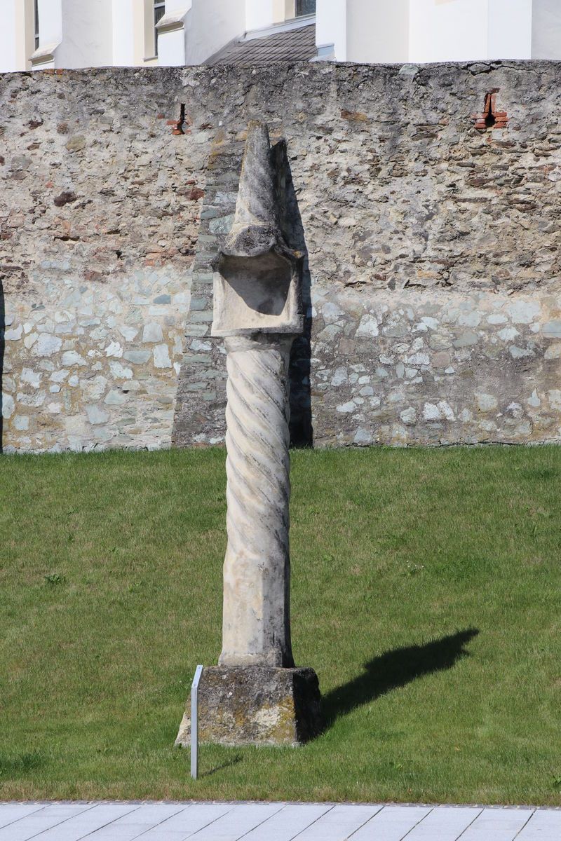 Eine alte Steinsäule mit Spiralrillen steht vor einer Steinmauer. Die Säule ist verwittert und wirft einen Schatten auf den Rasen.