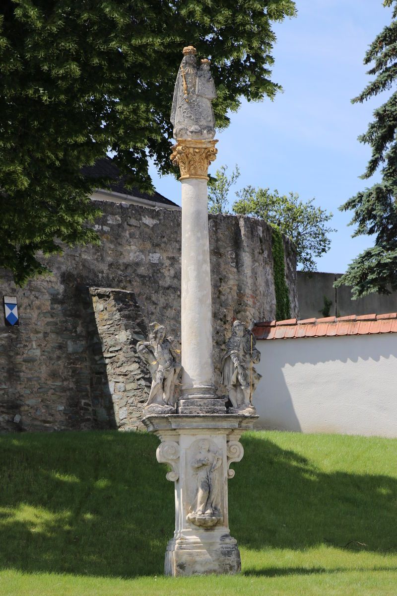 Eine klassische Steinsäule mit verzierten Schnitzereien steht in einem Garten, flankiert von zwei Statuen. Dahinter befindet sich eine Steinmauer mit einem blau-weißen Emblem.