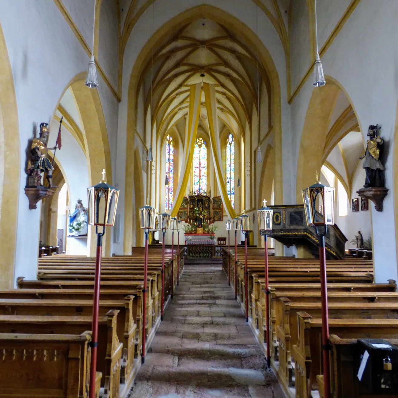 Der Innenraum einer Kirche mit hölzernen Kirchenbänken, gewölbten Decken und dekorativen Elementen wie Lampen und Statuen. Der Altar ist mit Blumen geschmückt, und Buntglasfenster sind sichtbar.