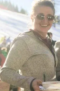 Eine Frau in einer grauen Kapuzenjacke lächelt und hält eine Schüssel in einer verschneiten Umgebung. Sie trägt eine Sonnenbrille und es befinden sich Menschen im unscharfen Hintergrund.