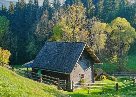 Ein Holzhaus mit Schieferdach steht auf einem grasbewachsenen Hügel, umgeben von einem Holzzaun. Im Hintergrund stehen Bäume.