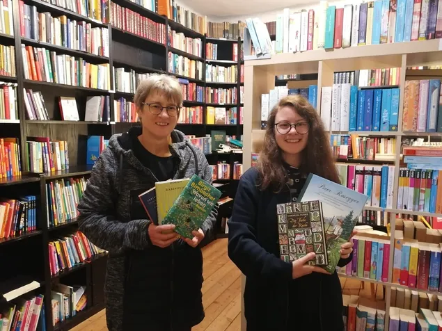 Zwei Frauen stehen in einem Buchladen mit Büchern in den Händen. Die Frau auf der linken Seite trägt eine Brille und eine Jacke, während die Frau auf der rechten Seite eine Brille und einen schwarzen Mantel trägt. Hinter ihnen befinden sich Regale voller Bücher.
