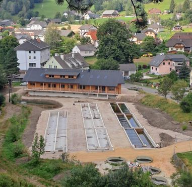 Luftaufnahme eines kleinen Alpendorfs, das eine Baustelle mit großen Betonwassertanks und einem modernen Gebäude im Bau zeigt.