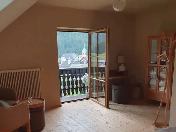 Ein Wohnzimmer mit Holzboden, einer Glastür, die zu einem Balkon führt, einem Holzschrank, einer Lampe, einer Couch und einem Heizkörper.