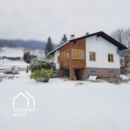 Eine schneebedeckte Landschaft mit einem Holzhaus mit gelb gerahmten Fenstern. Ein kleiner Busch und ein Stuhl stehen vor dem Haus.