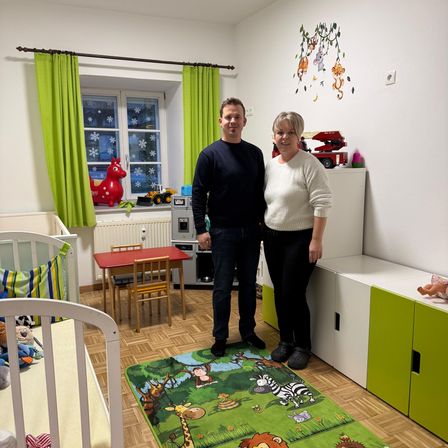 Ein Mann und eine Frau stehen in einem Kinderzimmer mit einem Teppich, Kinderbetten, einem Tisch, Stühlern und Spielzeug.