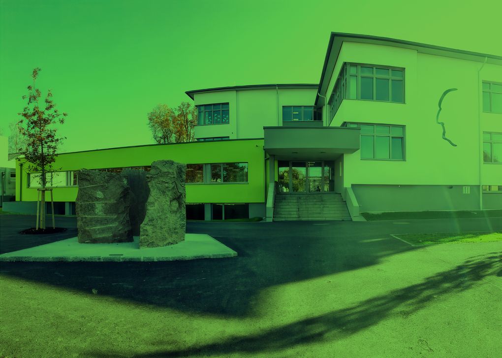  Abschnitt Bild: Ein modernes Gebäude mit grünen Wänden und großen Felsskulpturen davor. Der Eingang hat eine Treppe und eine Glastür.