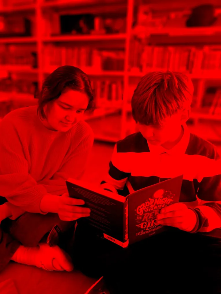 Abschnittsbild: Ein junges Mädchen und ein Junge sitzen auf dem Boden in einer Bibliothek und lesen ein Buch mit dem Titel 'Gute Nacht Geschichten für Rebellinnen'.