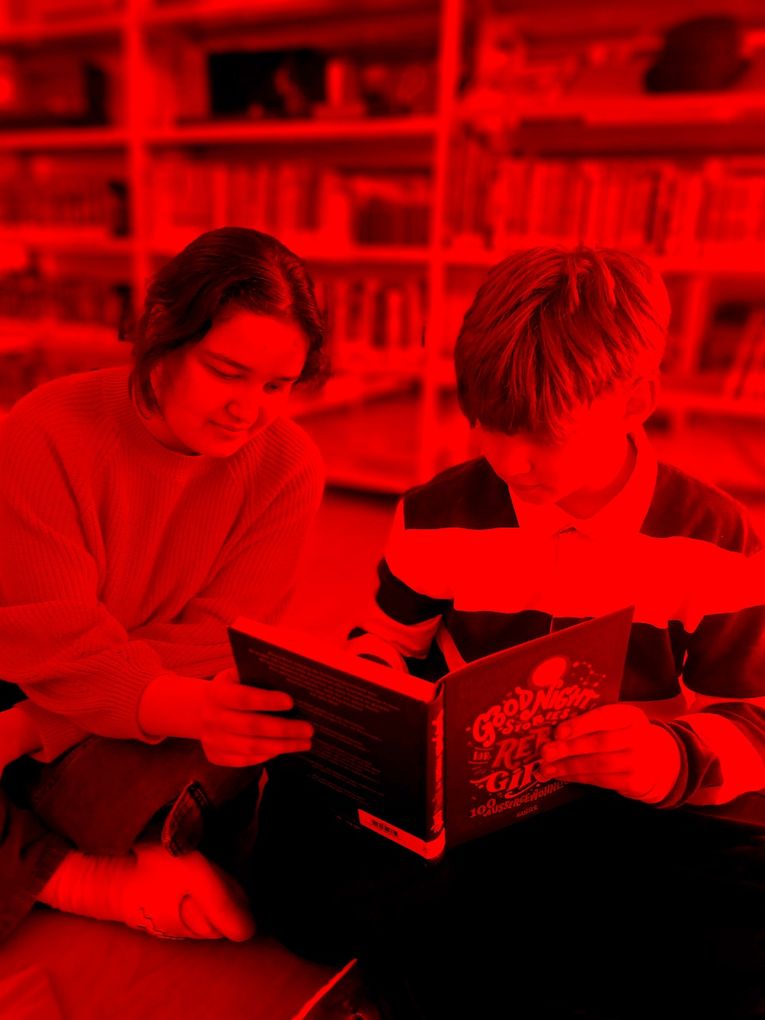 Abschnittsbild: Ein junges Mädchen und ein Junge sitzen auf dem Boden in einer Bibliothek und lesen ein Buch mit dem Titel 'Gute Nacht Geschichten für Rebellinnen'.