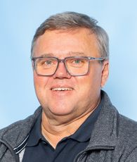 Ein lächelnder Mann mit grauem Haar und Brille steht vor einem blauen Hintergrund. Er trägt eine graue Jacke über einem schwarzen Hemd.