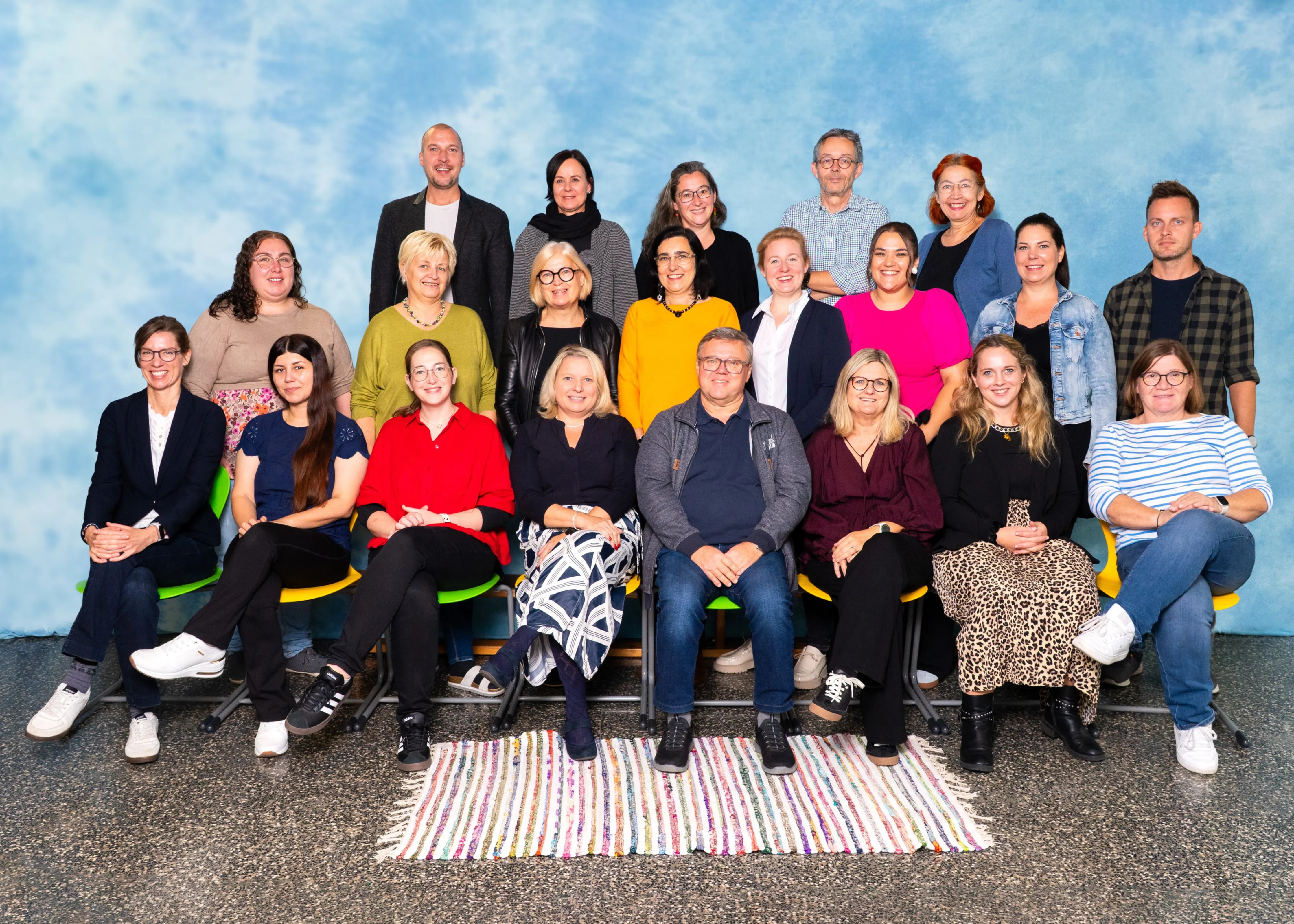 Eine Gruppe von Menschen in verschiedenen Kleidungsstücken und Accessoires posiert für ein Foto vor einem blauen Hintergrund mit Wolken.