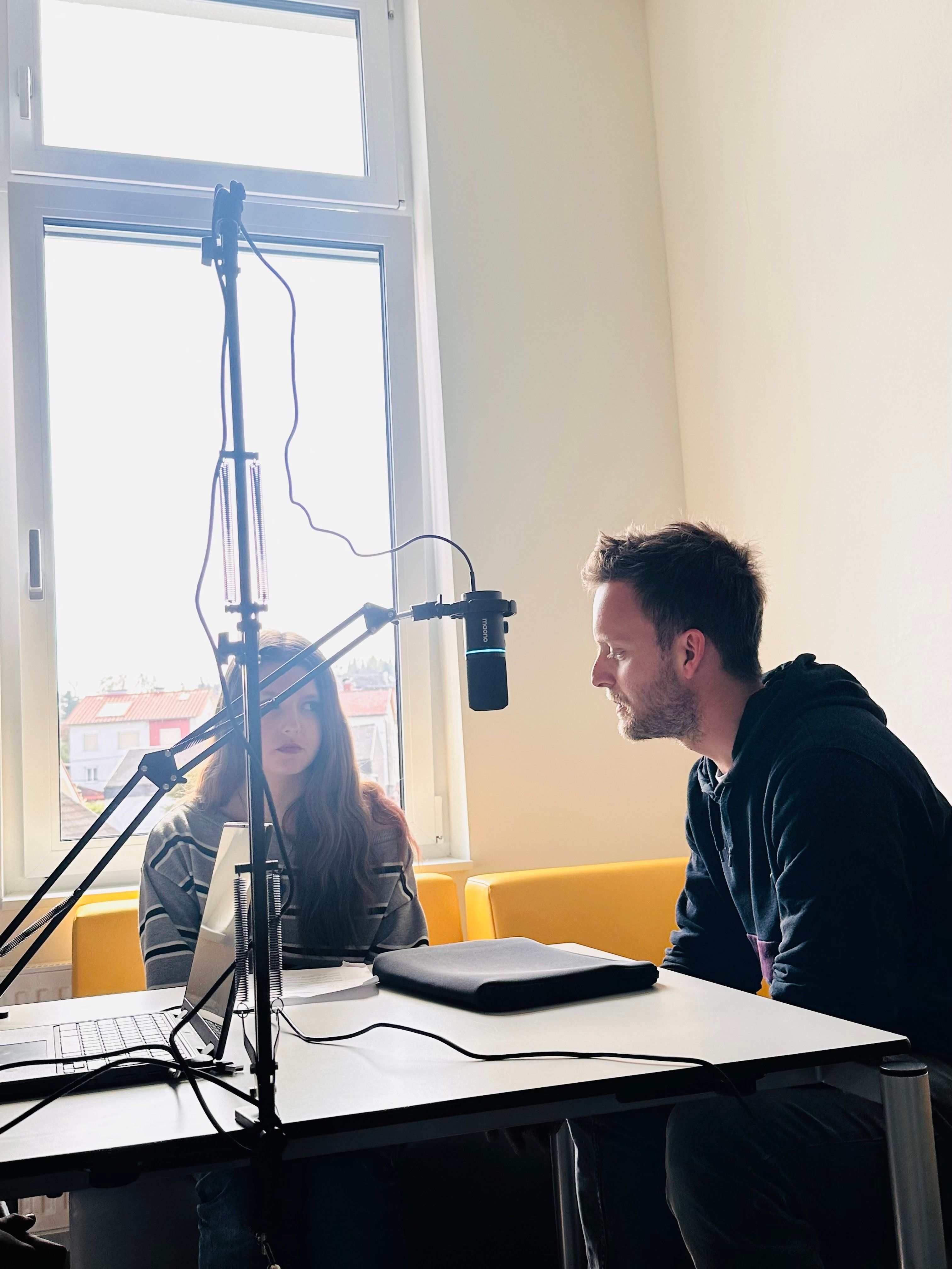 Ein Mann und eine Frau sitzen vor einem Mikrofon, möglicherweise bei einer Podcast-Aufnahme. Die Frau schaut auf den Laptop auf dem Tisch, während der Mann sie ansieht.