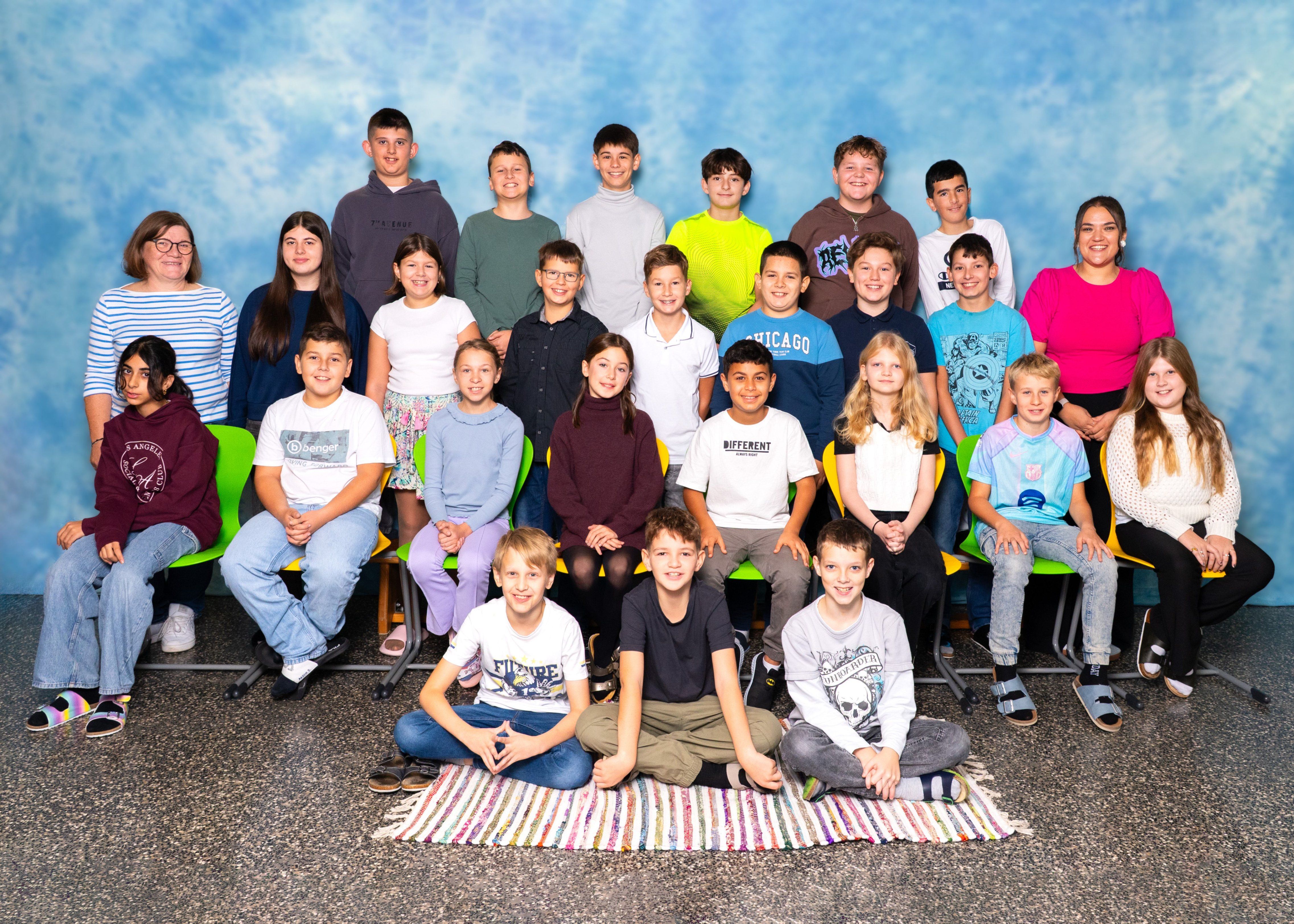 Eine Gruppe von Kindern und Jugendlichen posiert für ein Foto in einem Klassenzimmer mit blauem Hintergrund. Sie sitzen auf Stühlen und auf dem Boden, einige in lässiger Kleidung.