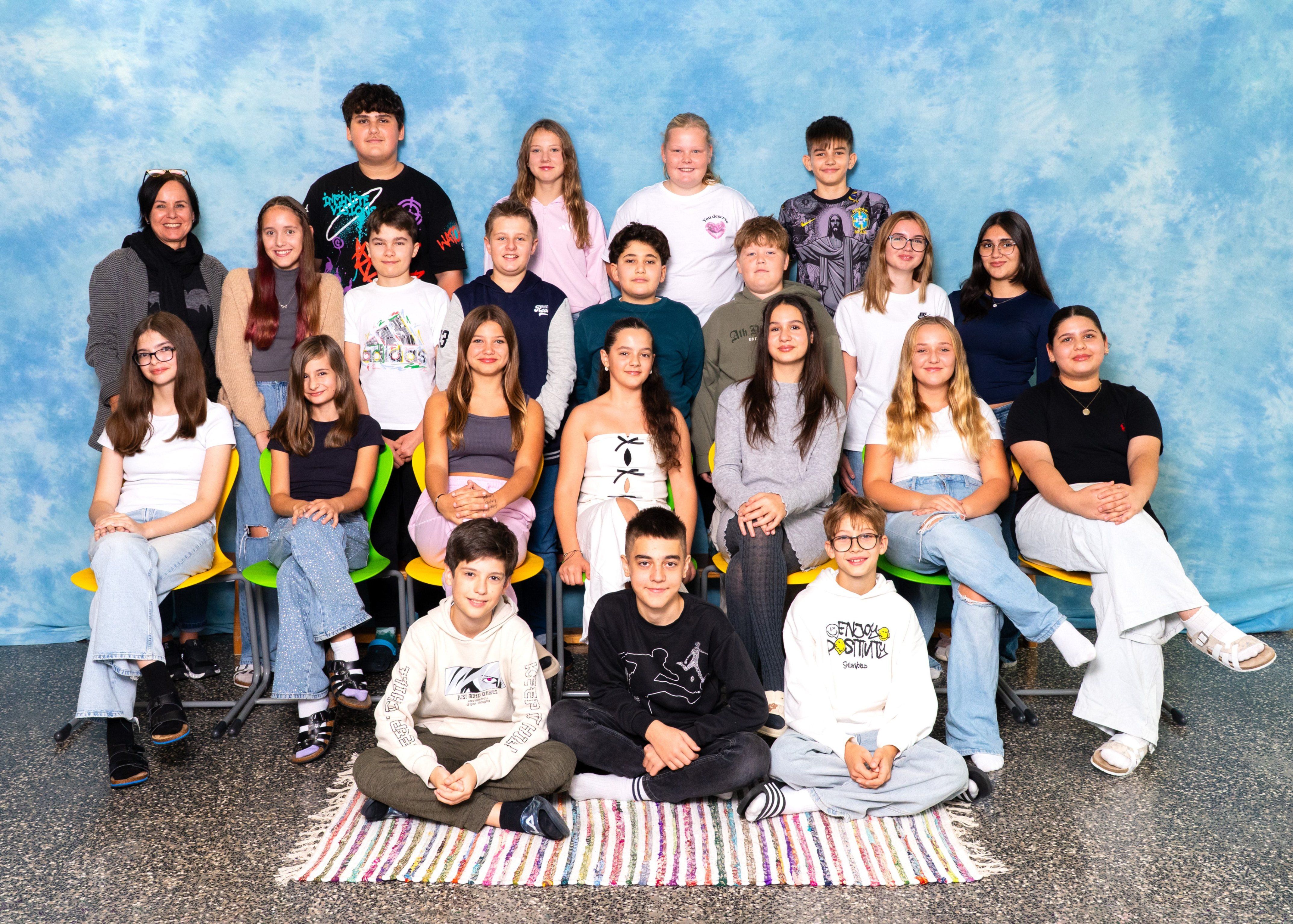 Eine Gruppe junger Menschen, einige sitzend und einige stehend, posiert für ein Foto vor einem blauen Hintergrund. Sie tragen Freizeitkleidung und scheinen sich in einem Studio zu befinden.