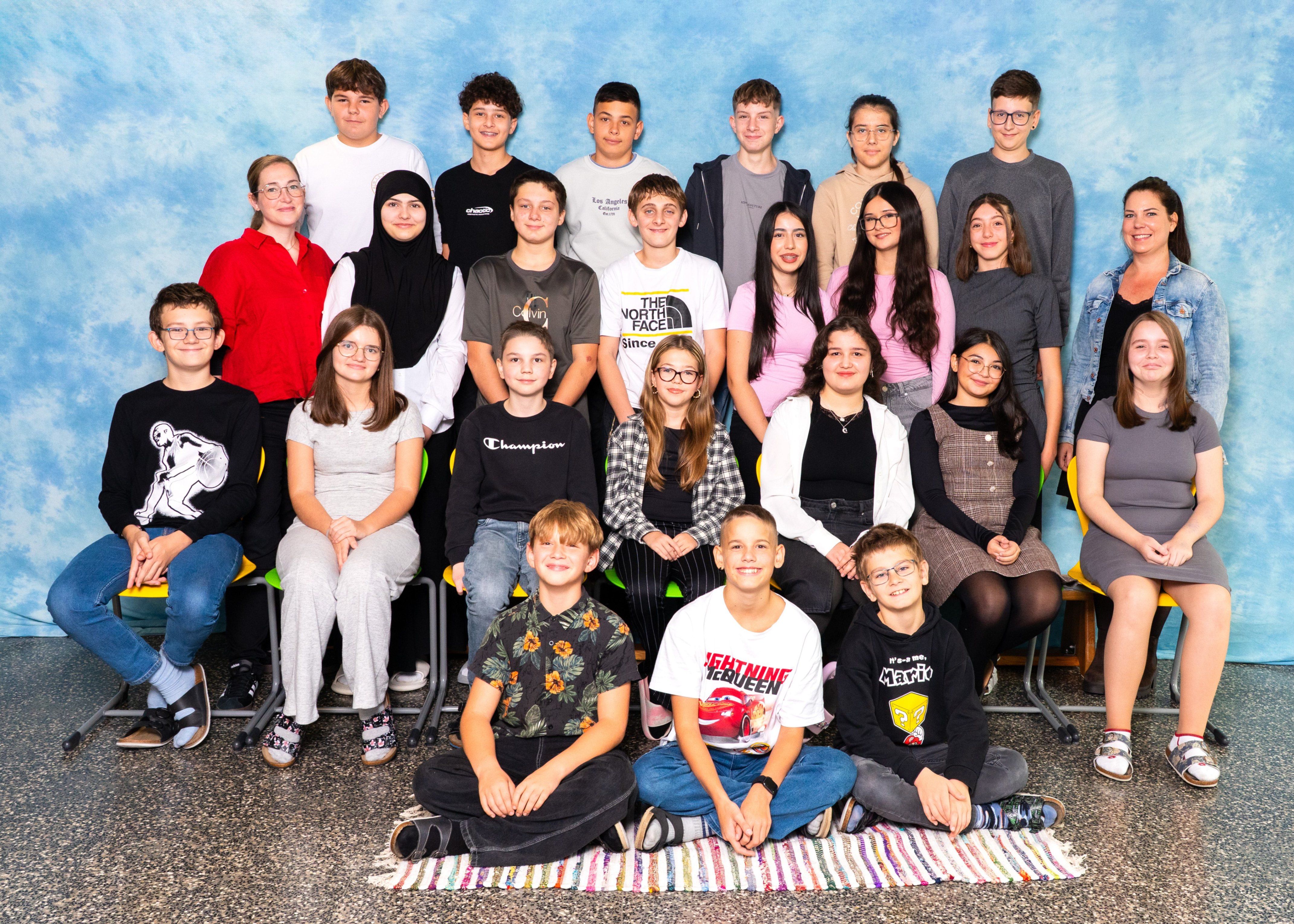 Eine Gruppe von Schülern, Jungen und Mädchen, posiert für ein Foto in einem Klassenzimmer. Sie sitzen und stehen im Halbkreis vor einem blauen Hintergrund.