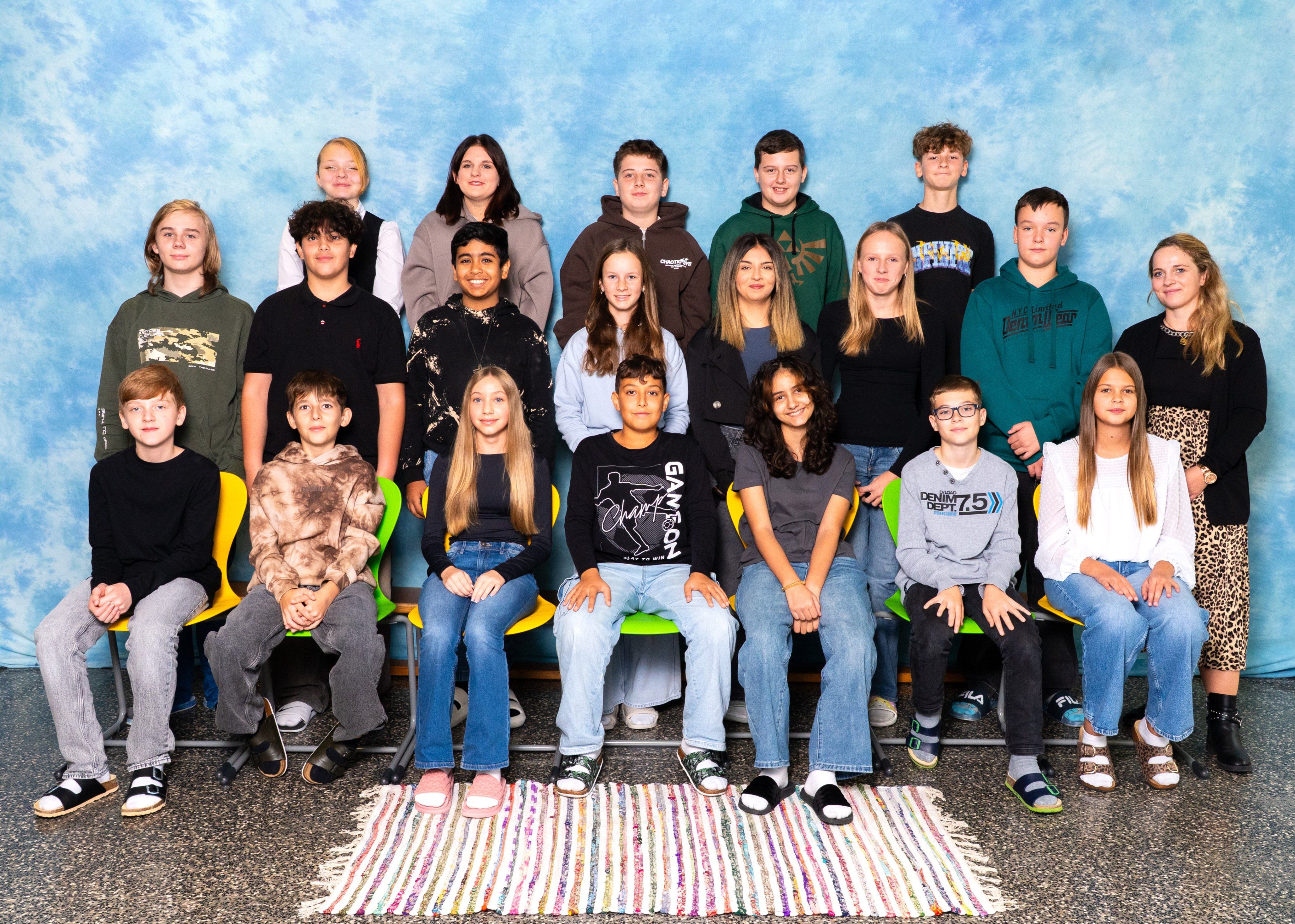 Eine Gruppe junger Menschen, darunter Jungen und Mädchen, posiert für ein Foto vor einem blauen Hintergrund. Sie sitzen auf grünen Stühlen, und einige stehen.