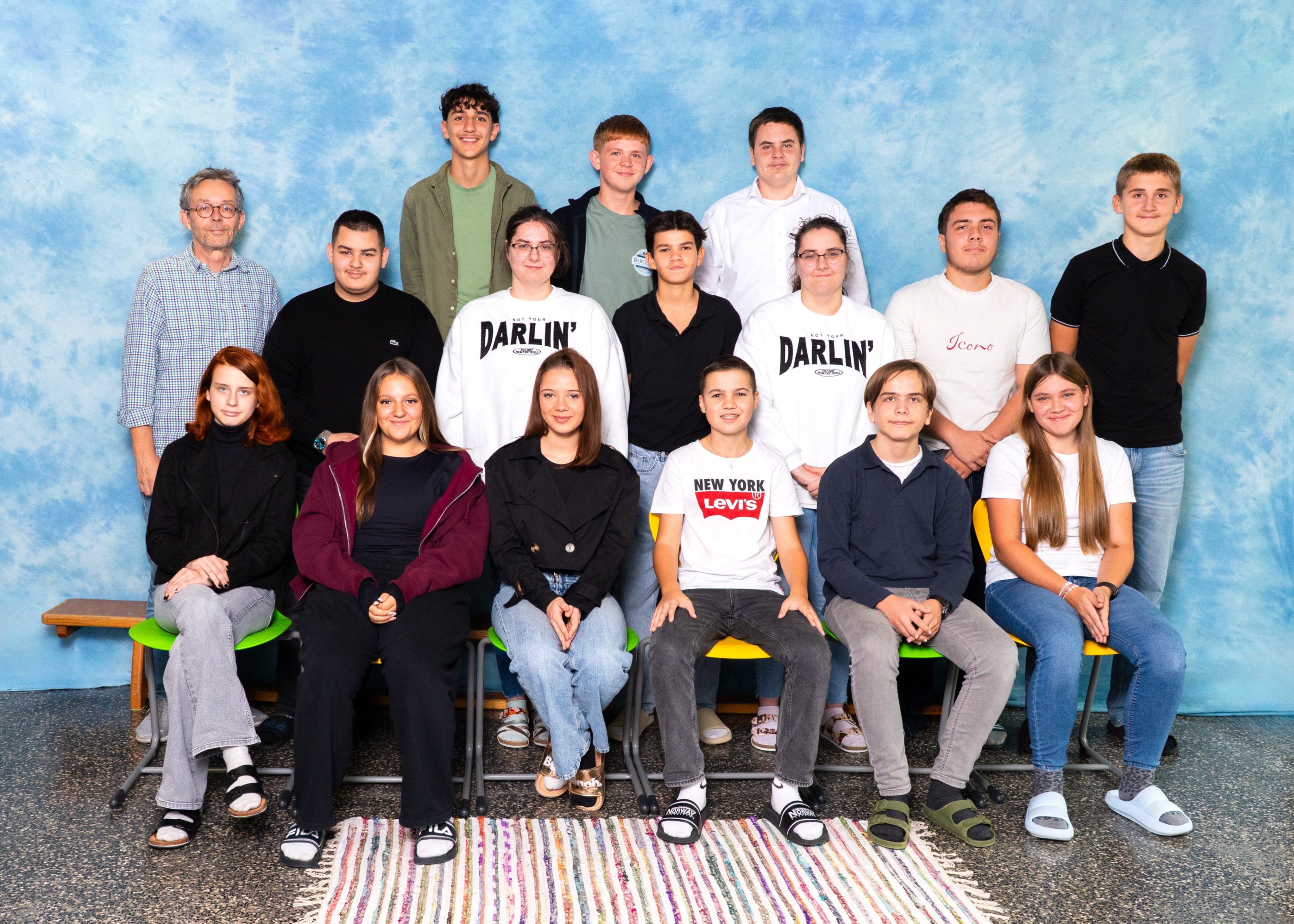 Eine Gruppe junger Menschen und Erwachsene posiert für ein Foto vor einem blauen Hintergrund. Sie sitzen auf Stühlen, einige tragen Brillen, Turnschuhe und verschiedene Kleidungsstücke.