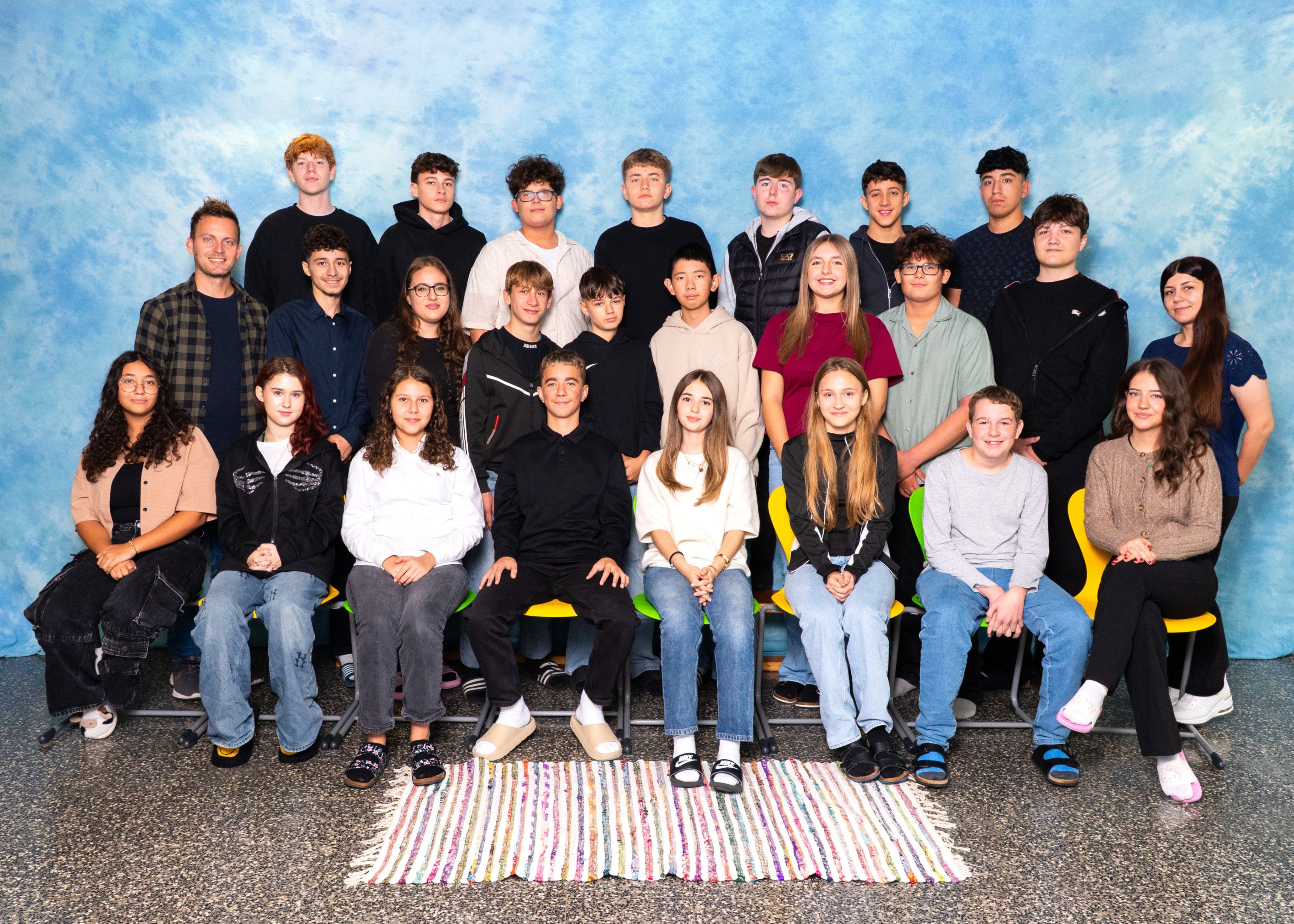 Eine Gruppe junger Menschen, sowohl Jungen als auch Mädchen, sitzen auf Stühlen und stehen vor einem blauen Hintergrund. Sie tragen alle Freizeitkleidung und scheinen für ein Foto zu posieren.