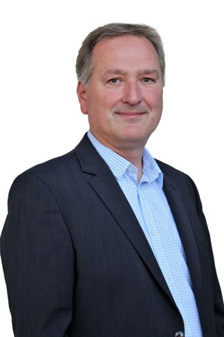 Ein Mann in einem blauen Hemd und einem dunklen Blazer steht lächelnd vor einem weißen Hintergrund.