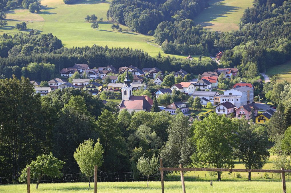  Abschnitt Bild: Ein friedliches Dorf, eingebettet in einem Tal, umgeben von üppigen grünen Feldern und dichten Wäldern. Eine Kirche mit einem Turm steht prominent im Zentrum.