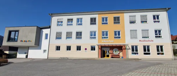 Die Vorderseite einer Volksschule, einer öffentlichen Grundschule in Deutschland, zeigt eine gelbe und weiße Fassade mit Fenstern, einer Bank und einem runden Fenster über dem Eingang.