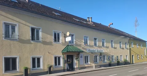 Ein beigefarbenes Gebäude mit mehreren Fenstern und einem grünen Vordach, mit einem Schild, das 'Gasthof Huber' und ein gelbes Schild über dem Eingang trägt. Zwei Topfpflanzen stehen vorne.