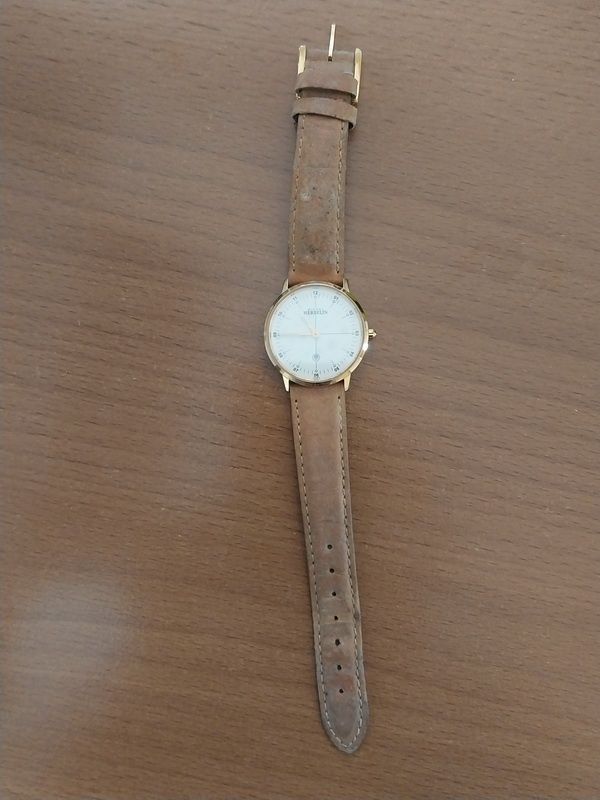 Eine goldfarbene Armbanduhr mit weißem Zifferblatt und braunem Lederarmband liegt auf einer Holzoberfläche.