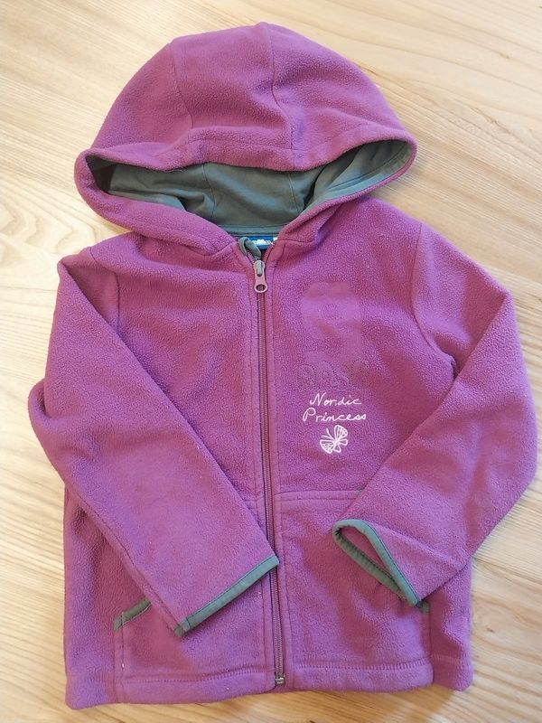 Ein lila Fleece-Hoodie mit Reißverschluss und einer Schmetterlings-Stickerei, die 'Nordic Princess' auf der Vorderseite sagt. Die Kapuze ist gefaltet und auf einer Holzoberfläche ausgestellt.