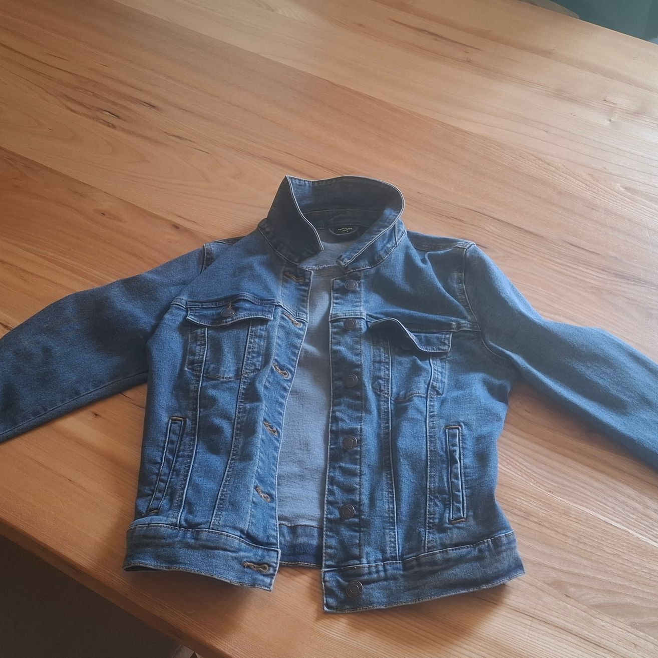 Eine Jeansjacke mit Kragen, Knöpfen und Taschen liegt flach auf einer Holzoberfläche.