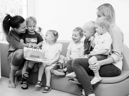 Eine Gruppe von Kindern sitzt auf einer Couch, lächelt und hört einer Geschichte zu, die von einer Frau vorgelesen wird. Zwei Frauen halten kleine Jungen auf ihren Knien.