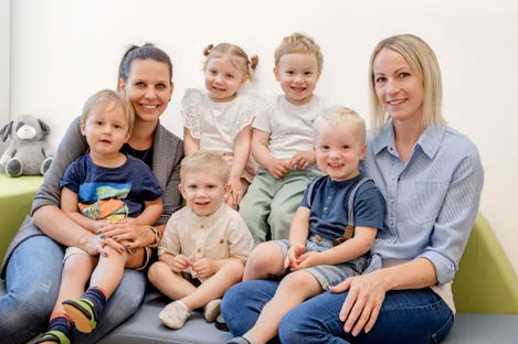 Eine Familie aus Frauen und Kindern sitzt zusammen auf einer Couch. Die Kinder lächeln und die Frauen lächeln zurück.