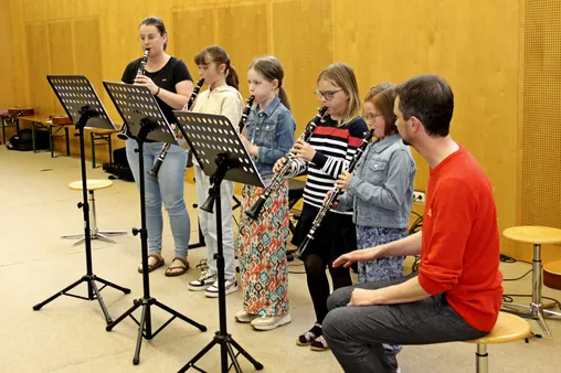 Ein Klarinettenlehrer mit fünf jungen Schülern. Sie spielen ihre Klarinetten. Die Schüler tragen Jacken, Brillen und Turnschuhe.