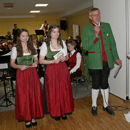 Zwei Frauen in traditioneller Kleidung stehen neben einem Mann, der ein Mikrofon hält. Dahinter spielt eine Band auf der Bühne, und Menschen sitzen und stehen im Hintergrund.
