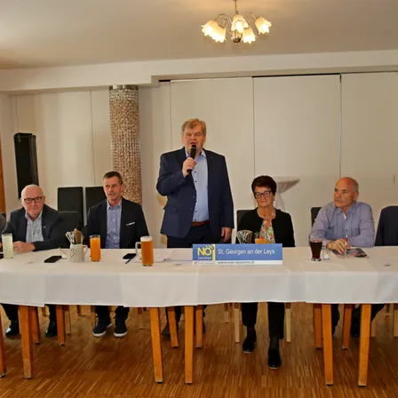 Eine Gruppe von Menschen sitzt um einen Tisch herum mit einem Mikrofon, einem Schild und Getränken. Ein Mann steht und spricht in das Mikrofon.