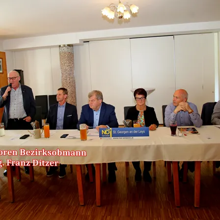 Eine Gruppe von Menschen sitzt bei einer Besprechung um einen Tisch. Eine Person spricht in ein Mikrofon. Auf dem Tisch stehen Gläser mit Getränken.