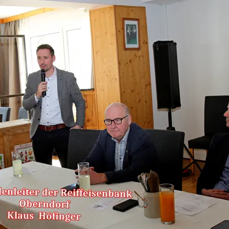 Klaus Hofinger, Geschäftsführer der Raiffeisenbank Oberndorf, sitzt an einem Tisch mit einem Glas Saft, während ein Mann hinter ihm steht und ein Mikrofon hält. Der Tisch hat verschiedene Gegenstände wie ein Glas, einen Becher und eine Mappe. Dahinter befindet sich eine Holzwand, ein Bilderrahmen und ein Lautsprecher.